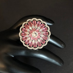 May include: A large, silver ring featuring a pink and white flower design. The ring is set with numerous small, clear stones and larger, pink stones.