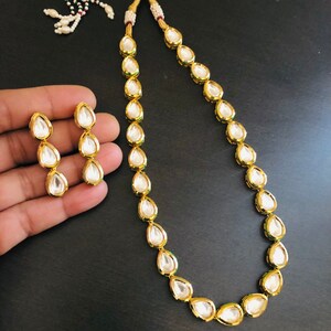 May include: A gold necklace and earrings set featuring teardrop-shaped white stones. The necklace is made of a gold chain with a series of teardrop-shaped stones.