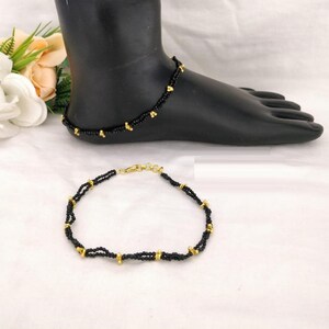 以下が含まれることがあります： 金色のアクセントが付いた黒いビーズのアンクレット2本。アンクレットは小さな黒いビーズで作られており、アンクレットに沿って金色のビーズが間隔をあけて配置されています。