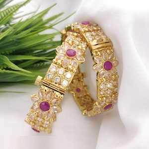May include: Two gold bangles with intricate floral designs. Each bangle is adorned with clear and pink gemstones.