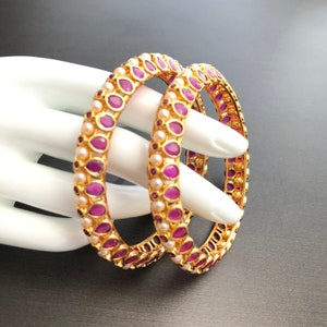 May include: Two gold bangles with purple and white stones. The bangles are stacked on top of each other.