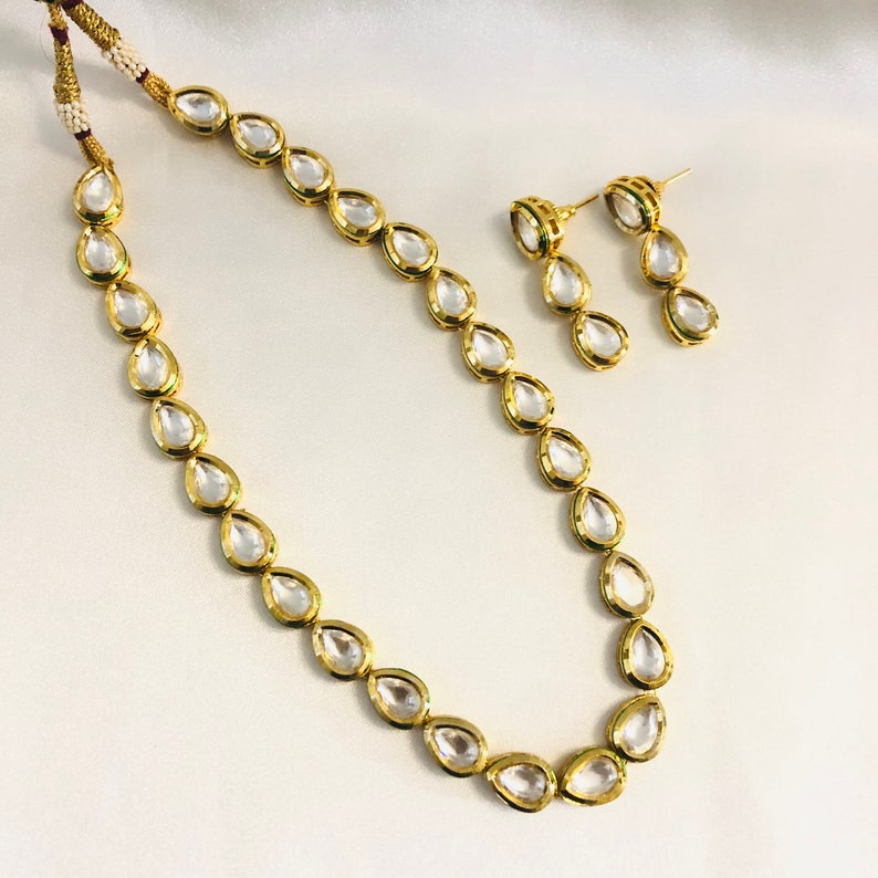 May include: A gold necklace and earrings set featuring clear teardrop-shaped stones. The necklace is made of a fine chain with a simple design. The earrings are drop style with three stones each.