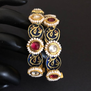 Puede incluir: Dos brazaletes de oro con esmalte azul y diseños florales intrincados. Cada brazalete está adornado con perlas blancas y piedras preciosas rojas y transparentes.