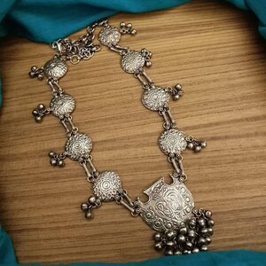 May include: A silver necklace with a large, ornate pendant and multiple smaller circular charms. The necklace is made of metal and has a delicate chain.