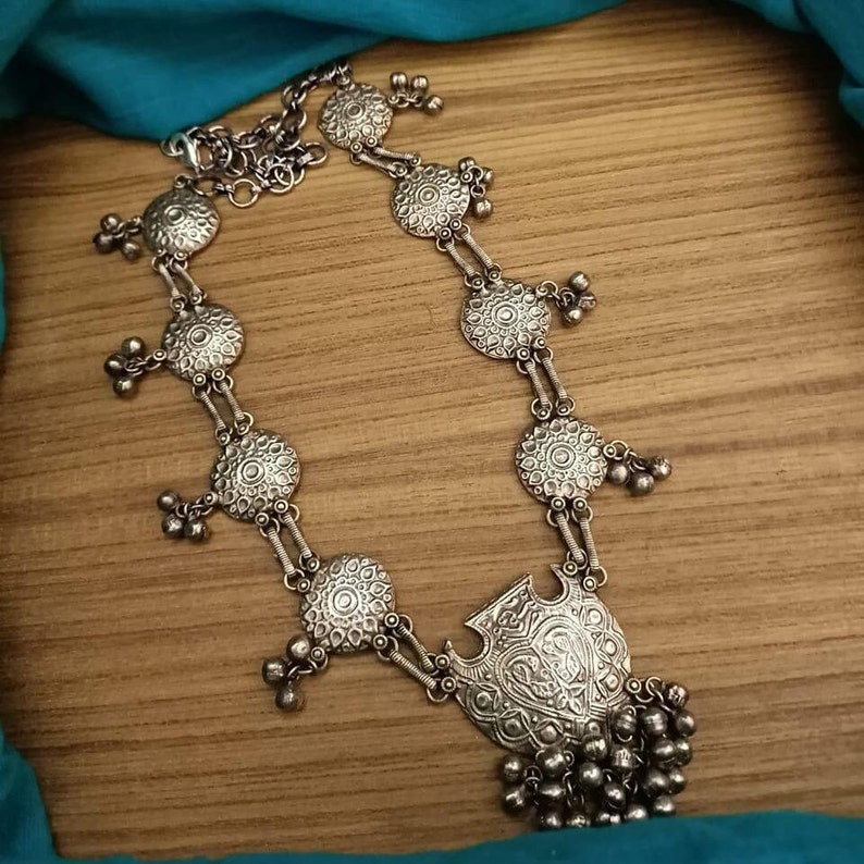 May include: A silver necklace with a large, ornate pendant and multiple smaller circular charms. The necklace is made of metal and has a delicate chain.