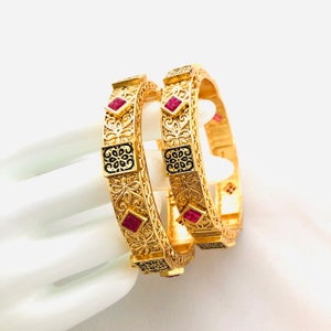 May include: Two gold bangles with intricate floral designs and pink gemstones. The bangles are adorned with black enamel accents.