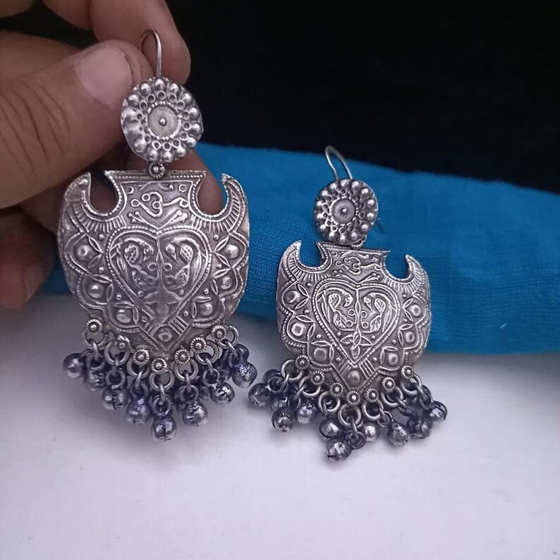 May include: A pair of silver earrings with a heart-shaped design and intricate details. The earrings have a dangling chain with small silver beads.