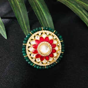 Könnte beinhalten: Ein runder, goldfarbener Anhänger mit einem roten und weißen Blumendesign. Die Blume hat eine weiße Mitte und ist von grünen Perlen umgeben.