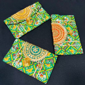May include: Three green and white patterned fabric envelopes with gold and colorful embellishments. Each envelope has a different colored embellishment, one is yellow, one is orange, and one is blue.