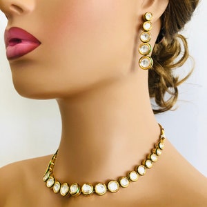 May include: A gold necklace and earrings set with round white stones and green accents. The necklace is a choker style with a row of stones.