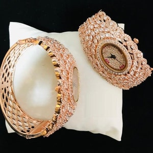 May include: A rose gold bangle watch with a white face and a diamond-like crystal design. The watch is on a white cushion.