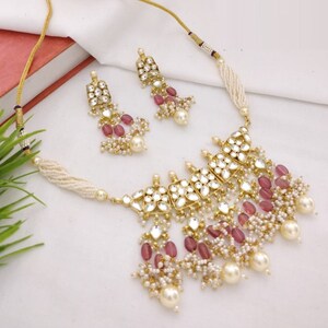May include: A gold-toned necklace and earring set. The necklace features a central design with clear, square-cut stones and pink beads, accented by dangling pearls. The earrings mirror the necklace design. The set is displayed on a white surface.