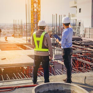 May include: Two construction workers wearing hard hats and safety vests on a building site. One points towards the structure while the other observes. A crane and the framework of a building are visible in the background.