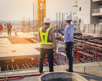 Puede incluir: Dos trabajadores de la construcción con cascos y chalecos de seguridad en una obra. Uno señala la estructura mientras el otro observa. Una grúa y la estructura de un edificio son visibles.