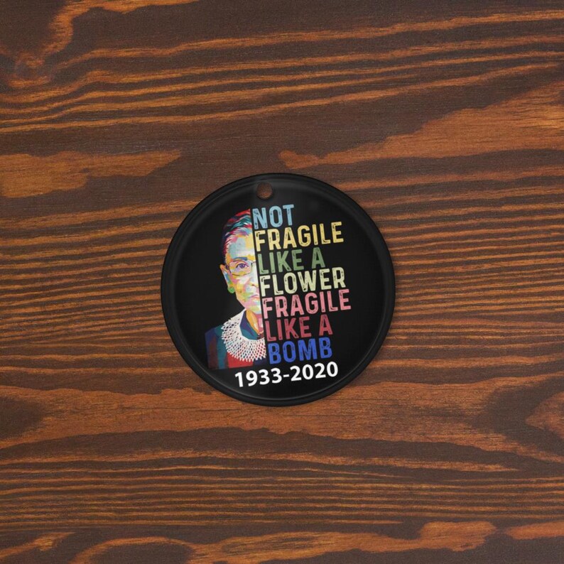 May include: A black circular ornament with a portrait and the text "NOT FRAGILE LIKE A FLOWER FRAGILE LIKE A BOMB 1933-2020". The design is colourful. It has a small hole at the top for hanging.