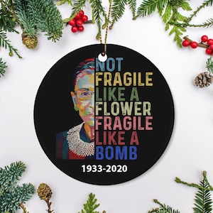 May include: A circular black ceramic ornament featuring a portrait of Ruth Bader Ginsburg. The ornament displays the text "NOT FRAGILE LIKE A FLOWER FRAGILE LIKE A BOMB" in various colours. The years 1933-2020 are printed at the bottom.