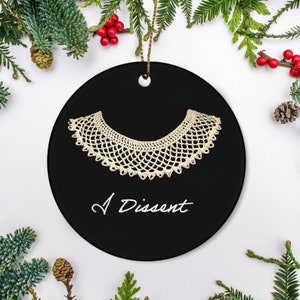 May include: Black circular ceramic ornament with a cream-coloured lace collar design and the words "I Dissent" in white script. The ornament has a gold string for hanging, surrounded by festive greenery.