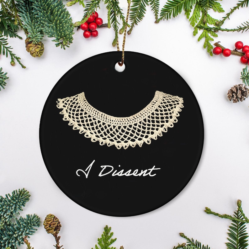May include: Black circular ceramic ornament with a cream-coloured lace collar design and the words "I Dissent" in white script. The ornament has a gold string for hanging, surrounded by festive greenery.