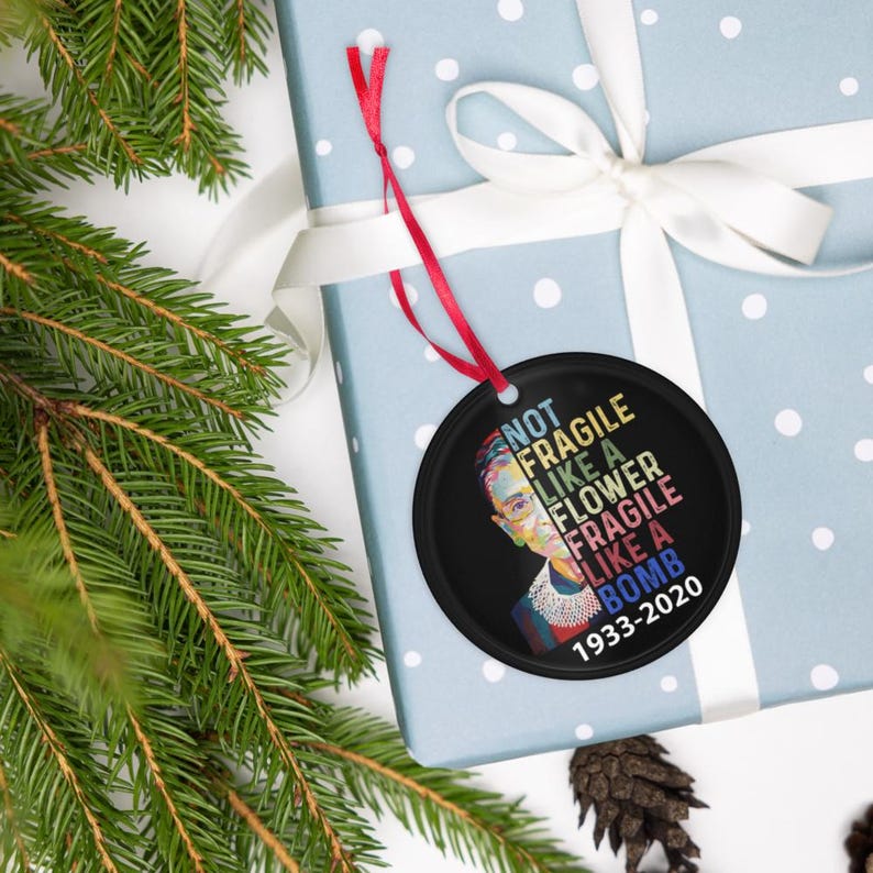 May include: A black circular Christmas ornament featuring a portrait of Ruth Bader Ginsburg and the text "NOT FRAGILE LIKE A FLOWER FRAGILE LIKE A BOMB 1933-2020". The ornament hangs from a red ribbon, on a light blue gift box.