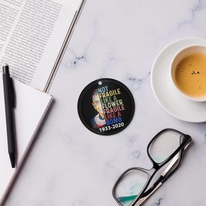 May include: A black circular ornament featuring the text "NOT FRAGILE LIKE A FLOWER FRAGILE LIKE A BOMB 1933-2020" in multiple colours, and a portrait of a woman. The ornament has a hole at the top for hanging.