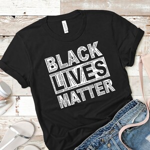 May include: Black t-shirt with the words "BLACK LIVES MATTER" in white, with names of people in the letters. The shirt is folded, with a pair of blue jeans, a pink purse, and white headphones in the background.