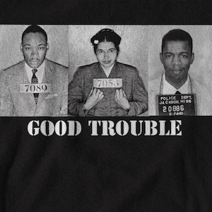 May include: A black and white graphic featuring three portraits in the style of mugshots of civil rights leaders. The words "GOOD TROUBLE" are printed in white block letters below the portraits. Each person holds a sign with a number.