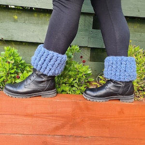 May include: A person wearing black leggings and black leather boots with blue knitted boot cuffs. The boot cuffs are a chunky knit design and are a light blue color. The boots are on a wooden surface.