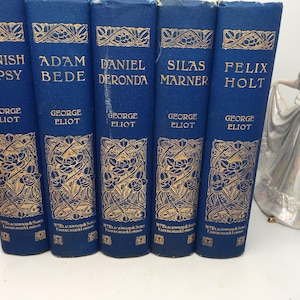 May include: A set of six blue hardcover books with gold lettering and decorative designs. The books are titled "Spanish Gypsy", "Adam Bede", "Daniel Deronda", "Silas Marner", and "Felix Holt". Each book is by George Eliot.
