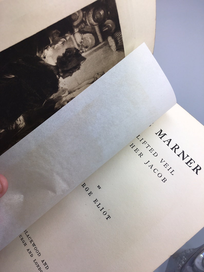 May include: A vintage book titled "Lifted Veil" by R.G.E. Elliot. The book is open to a page with the title "The Marner" and the text "Lifted Veil" and "Her Jacob".