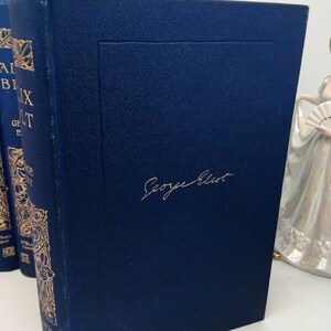May include: A dark blue hardcover book with gold lettering and a gold decorative design on the spine. The book is titled "Adam Bede" by George Eliot. The author's name is written in gold cursive on the front cover.