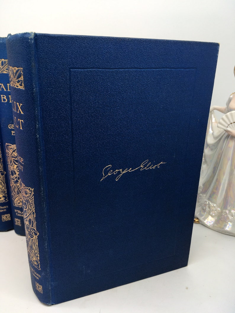 May include: A dark blue hardcover book with gold lettering and a gold decorative design on the spine. The book is titled "Adam Bede" by George Eliot. The author's name is written in gold cursive on the front cover.