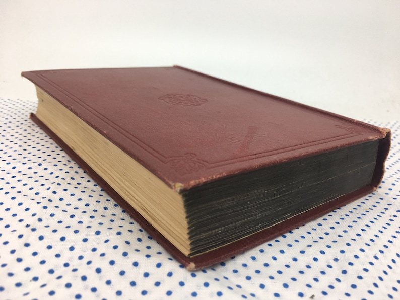 K&ouml;nnte beinhalten: Ein burgunderfarbenes Hardcover-Buch mit einem goldenen Design auf dem vorderen Umschlag. Das Buch ist geschlossen und liegt auf einem wei&szlig;en Stoff mit blauen Punkten.