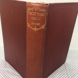 K&ouml;nnte beinhalten: Ein rotes Hardcover-Buch mit goldenen Lettern auf dem R&uuml;cken. Der Titel lautet "Onkel Toms H&uuml;tte" von Stowe.