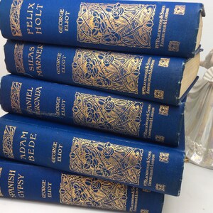 May include: A stack of seven blue hardcover books with gold lettering and decorative designs. The books are titled "Spanish Gypsy", "Adam Bede", "Daniel Deronda", "Silas Marner", "Felix Holt", and "The Mill on the Floss".
