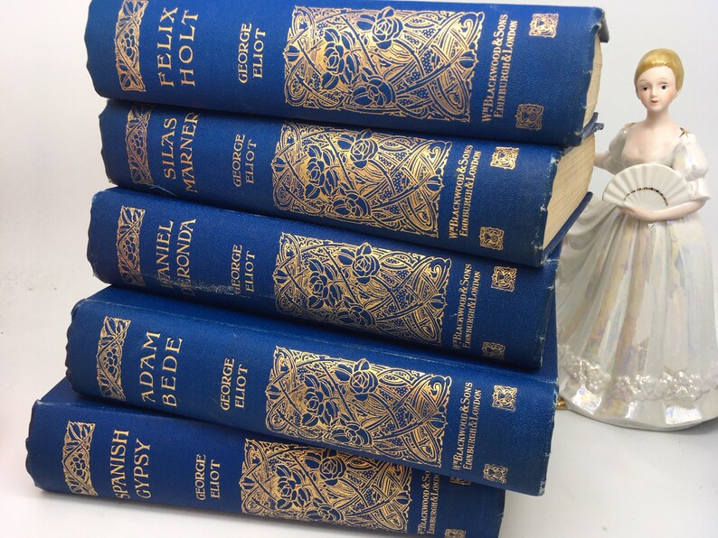 May include: A stack of seven blue hardcover books with gold lettering and decorative designs. The books are titled "Spanish Gypsy", "Adam Bede", "Daniel Deronda", "Silas Marner", "Felix Holt", and "The Mill on the Floss".