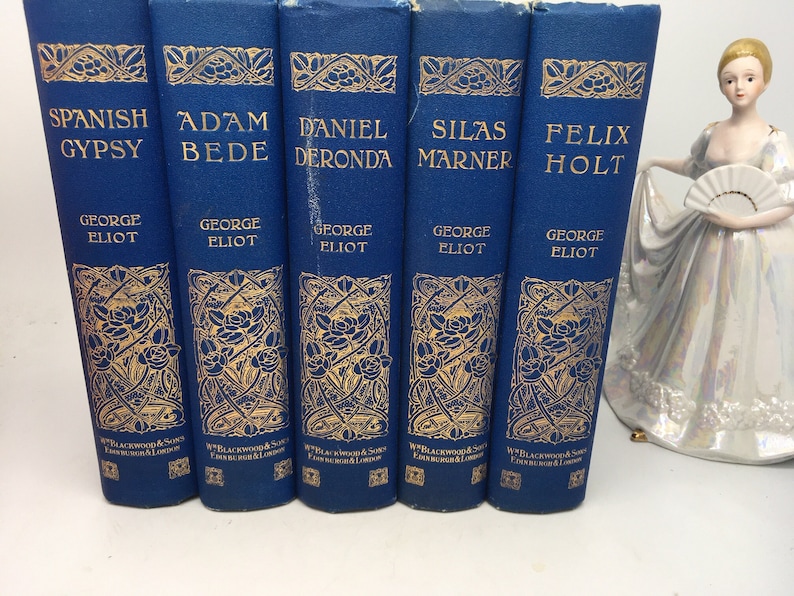 May include: A set of six blue hardcover books with gold lettering and decorative designs. The books are titled "Spanish Gypsy", "Adam Bede", "Daniel Deronda", "Silas Marner", and "Felix Holt". Each book is by George Eliot.