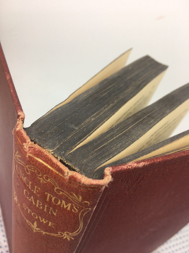 K&ouml;nnte beinhalten: Ein antikes Hardcover-Buch mit rotem Einband und goldenen Lettern. Der Titel lautet "Onkel Toms H&uuml;tte" von Harriet Beecher Stowe.