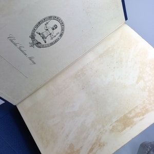 May include: An open book with a white cover and a blue spine. The book has a decorative seal on the front cover with a man holding a sword and a key. The text on the seal reads "Furth Fortune and Fill the Fetters". The text "Charles Scribner's Sons" is written on the cover.