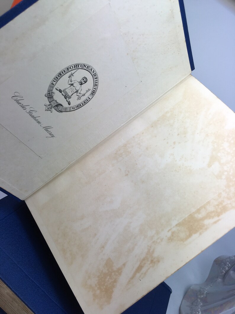 May include: An open book with a white cover and a blue spine. The book has a decorative seal on the front cover with a man holding a sword and a key. The text on the seal reads "Furth Fortune and Fill the Fetters". The text "Charles Scribner's Sons" is written on the cover.