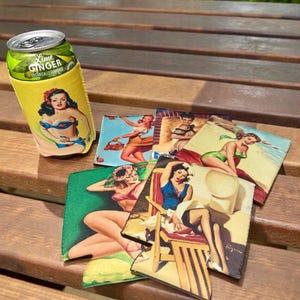 May include: A yellow can cooler with a vintage pin-up girl design, holding a can of Lime Ginger. Several matching coasters with different pin-up girl illustrations are arranged on a wooden surface. The coasters feature women in swimwear.