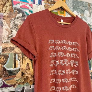 K&ouml;nnte beinhalten: Ein rostfarbenes Kurzarm-T-Shirt mit einem sich wiederholenden wei&szlig;en Grafikdruck von G&uuml;rteltieren, die in horizontalen Reihen angeordnet sind. Das T-Shirt hat einen Rundhalsausschnitt und h&auml;ngt an einem Holzkleiderb&uuml;gel mit hochgekrempelten &Auml;rmeln.