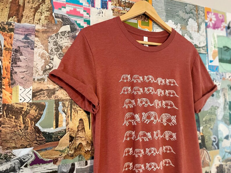 K&ouml;nnte beinhalten: Ein rostfarbenes Kurzarm-T-Shirt mit einem sich wiederholenden wei&szlig;en Grafikdruck von G&uuml;rteltieren, die in horizontalen Reihen angeordnet sind. Das T-Shirt hat einen Rundhalsausschnitt und h&auml;ngt an einem Holzkleiderb&uuml;gel mit hochgekrempelten &Auml;rmeln.