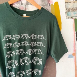 K&ouml;nnte beinhalten: Ein dunkelwaldgr&uuml;nes Kurzarm-T-Shirt mit einem sich wiederholenden wei&szlig;en Grafikdruck von G&uuml;rteltieren, die in horizontalen Reihen auf der Vorderseite angeordnet sind. Das T-Shirt hat einen klassischen Rundhalsausschnitt und h&auml;ngt an einem Holzkleiderb&uuml;gel.