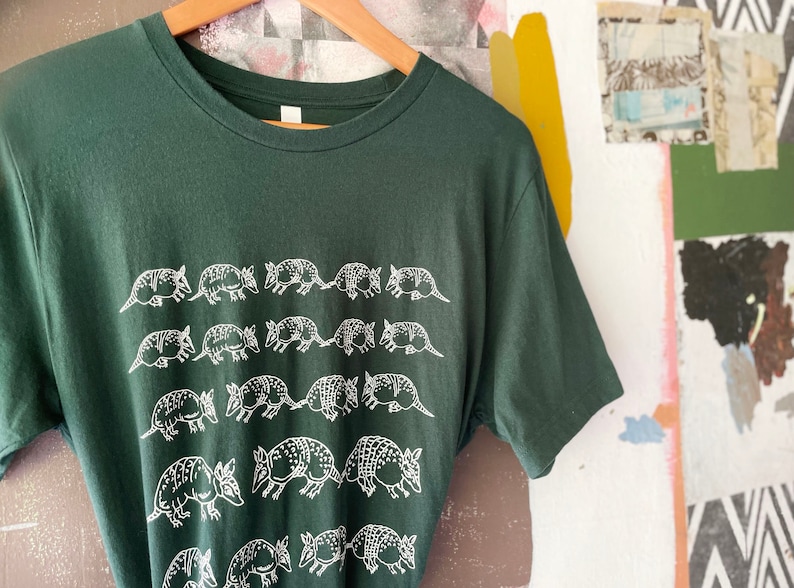 K&ouml;nnte beinhalten: Ein dunkelwaldgr&uuml;nes Kurzarm-T-Shirt mit einem sich wiederholenden wei&szlig;en Grafikdruck von G&uuml;rteltieren, die in horizontalen Reihen auf der Vorderseite angeordnet sind. Das T-Shirt hat einen klassischen Rundhalsausschnitt und h&auml;ngt an einem Holzkleiderb&uuml;gel.