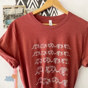 K&ouml;nnte beinhalten: Ein rostfarbenes, meliertes Kurzarm-T-Shirt mit einem wei&szlig;en Grafikdruck von mehreren G&uuml;rteltieren, die in Reihen angeordnet sind. Das T-Shirt hat einen Rundhalsausschnitt und wird auf einem Holzkleiderb&uuml;gel pr&auml;sentiert.