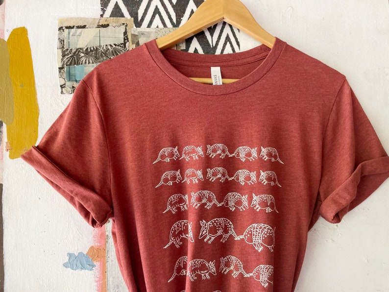 K&ouml;nnte beinhalten: Ein rostfarbenes, meliertes Kurzarm-T-Shirt mit einem wei&szlig;en Grafikdruck von mehreren G&uuml;rteltieren, die in Reihen angeordnet sind. Das T-Shirt hat einen Rundhalsausschnitt und wird auf einem Holzkleiderb&uuml;gel pr&auml;sentiert.