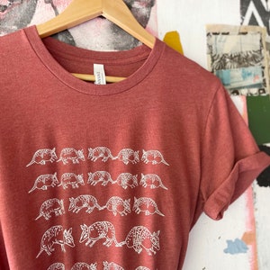 K&ouml;nnte beinhalten: Ein meliertes, terrakottarotes T-Shirt mit Rundhalsausschnitt und einem sich wiederholenden wei&szlig;en Linienmuster aus G&uuml;rteltieren, die in vier horizontalen Reihen angeordnet sind. Das T-Shirt h&auml;ngt an einem Holzkleiderb&uuml;gel und hat ein wei&szlig;es Etikett.