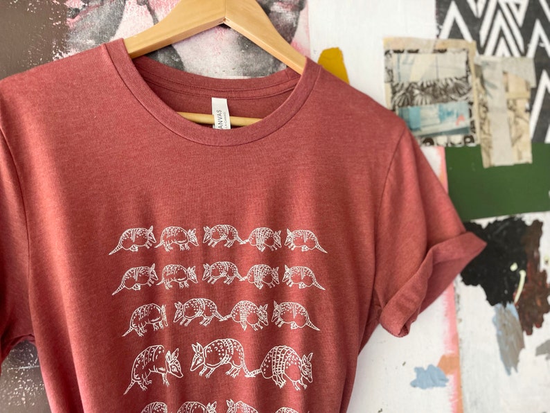 K&ouml;nnte beinhalten: Ein meliertes, terrakottarotes T-Shirt mit Rundhalsausschnitt und einem sich wiederholenden wei&szlig;en Linienmuster aus G&uuml;rteltieren, die in vier horizontalen Reihen angeordnet sind. Das T-Shirt h&auml;ngt an einem Holzkleiderb&uuml;gel und hat ein wei&szlig;es Etikett.