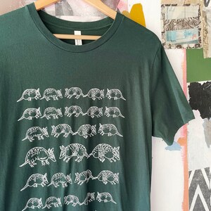 K&ouml;nnte beinhalten: Ein dunkelwaldgr&uuml;nes Kurzarm-T-Shirt mit einem sich wiederholenden wei&szlig;en Grafikdruck von G&uuml;rteltieren, die in horizontalen Reihen auf der Vorderseite angeordnet sind. Das T-Shirt hat einen Rundhalsausschnitt und h&auml;ngt an einem Holzkleiderb&uuml;gel.
