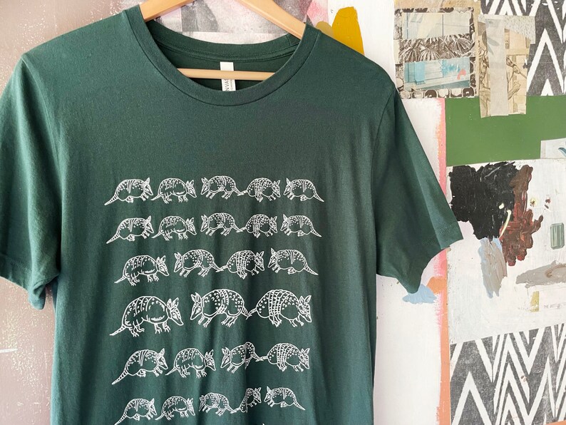 K&ouml;nnte beinhalten: Ein dunkelwaldgr&uuml;nes Kurzarm-T-Shirt mit einem sich wiederholenden wei&szlig;en Grafikdruck von G&uuml;rteltieren, die in horizontalen Reihen auf der Vorderseite angeordnet sind. Das T-Shirt hat einen Rundhalsausschnitt und h&auml;ngt an einem Holzkleiderb&uuml;gel.